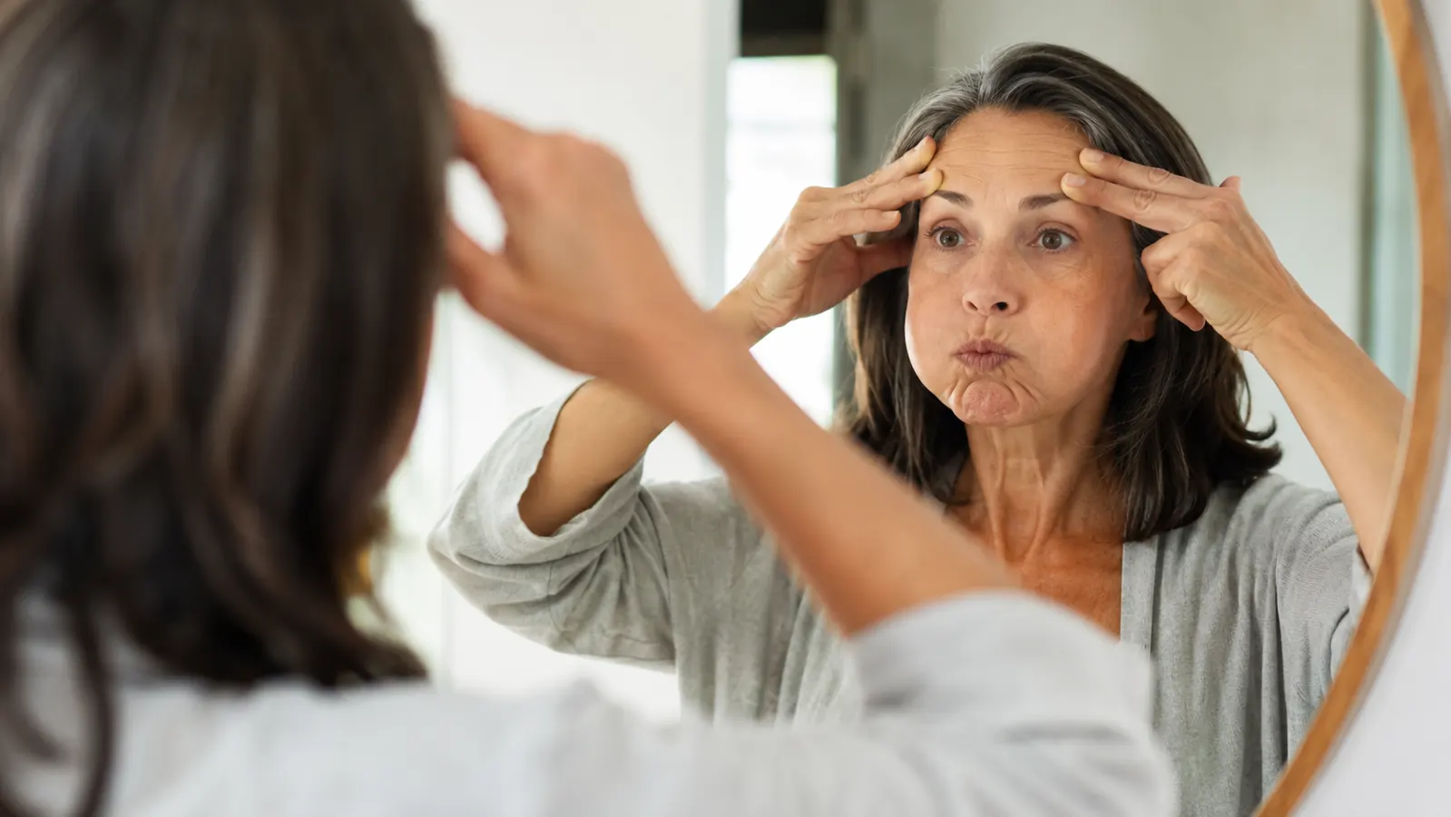 Woman doing a face yoga cheek-lift exercise in front of a bathroom mirror, morning light