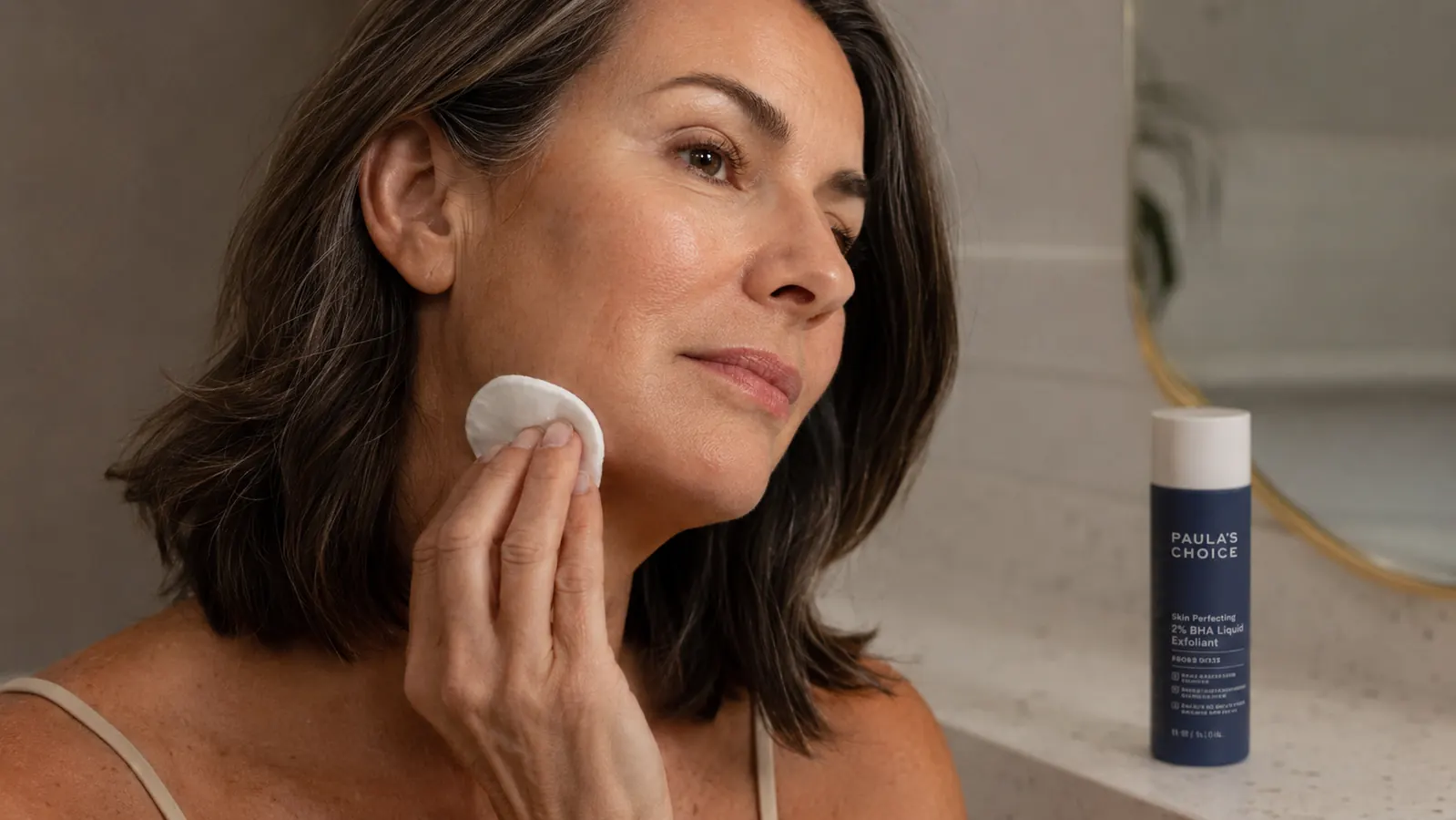 A hand pressing a cotton round soaked in liquid exfoliant along the jaw and chin, bottle of Paula's Choice 2% BHA visible on the counter
